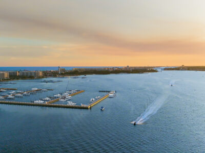Aerial view of a marina with numerous yachts and boats docked, surrounded by water, with a cityscape in the background under a sunset sky in Fort Walton Beach.