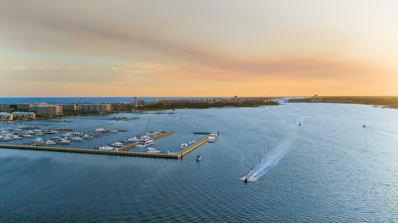 Aerial view of a marina with numerous yachts and boats docked, surrounded by water, with a cityscape in the background under a sunset sky in Fort Walton Beach.