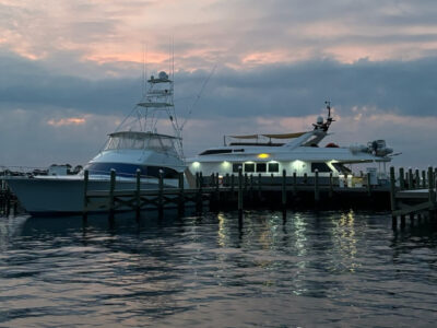 A serene marina at dusk in Fort Walton Beach, showcasing two boats moored at a wooden dock with a vibrant sunset reflecting on the calm water.