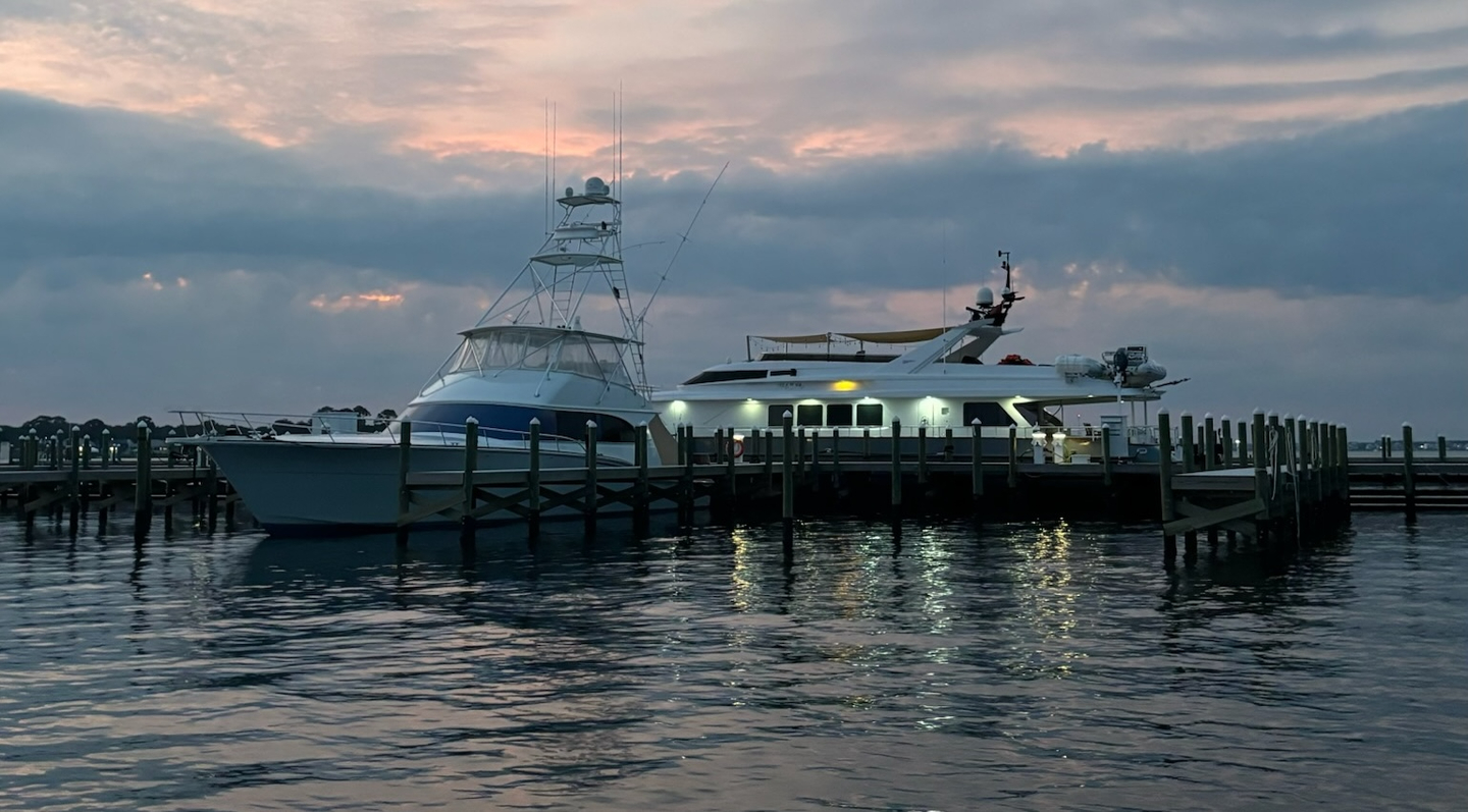A serene marina at dusk in Fort Walton Beach, showcasing two boats moored at a wooden dock with a vibrant sunset reflecting on the calm water.