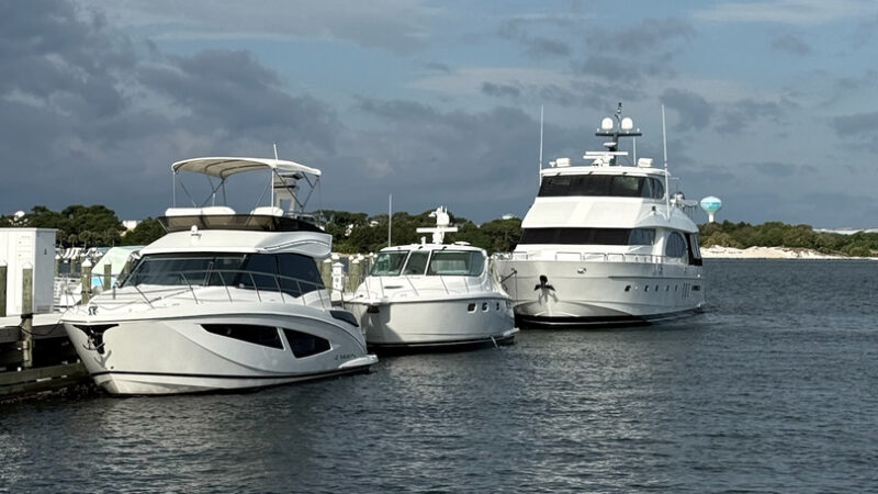 Luxury yachts docked at marina transient slips under partly cloudy skies in Fort Walton Beach, with calm waters and a scenic backdrop of lush greenery and distant water views.