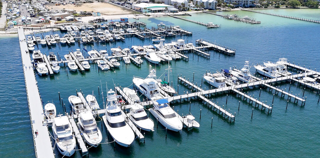 Aerial view of a bustling marina in Fort Walton Beach, showcasing numerous docked yachts and boats with clear blue waters and surrounding waterfront buildings.